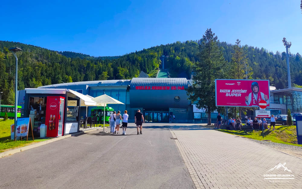 Entrance to a mountain ski-lift area with a blue station building, people walking, and outdoor cafe seating under umbrellas. A large pink billboard is on the right.
