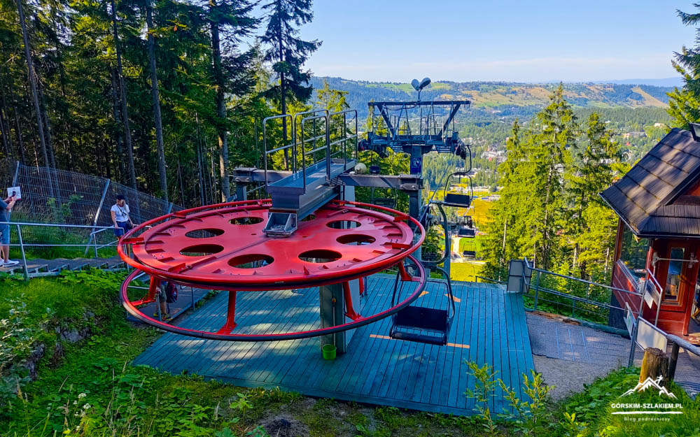 Red circular cable-car wheel on a blue platform with a forested mountain valley in the background, empty chairs nearby.