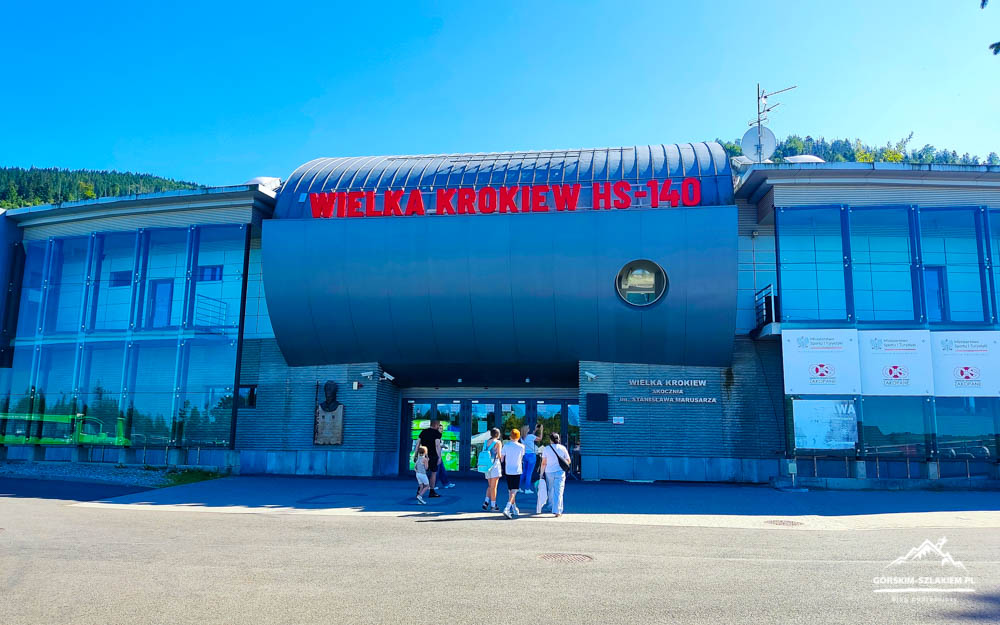 Entrance to the Wielka Krokiew HS-140 sport facility with a curved canopy and a group of people walking toward the doors