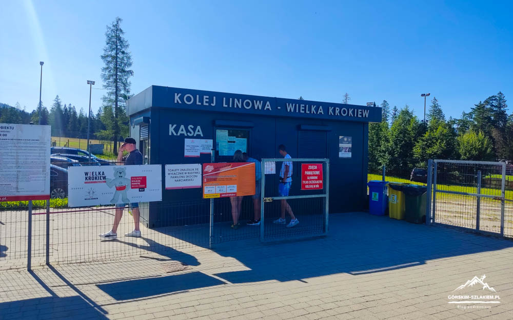 Blue ticket booth labeled KASA at Kolej Linowa – Wielka Krokiew, with people in front buying tickets and signs posted on a fence nearby on a sunny day.