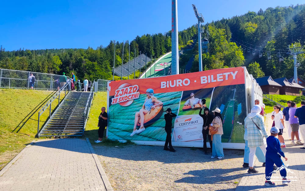 Ticket booth tent with Polish 'BIURO BILETY' at an outdoor park; people walk nearby with a ski-jump slope in the background.