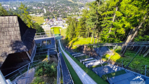 A long outdoor ramp descending a forested hillside, with metal rails and green turf, overlooking a town in the valley.
