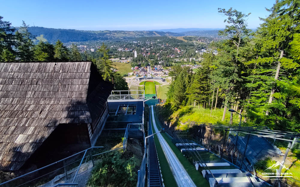 Ski jumping ramp and wooden lodge on a forested hillside, with a valley town and blue sky in the background.