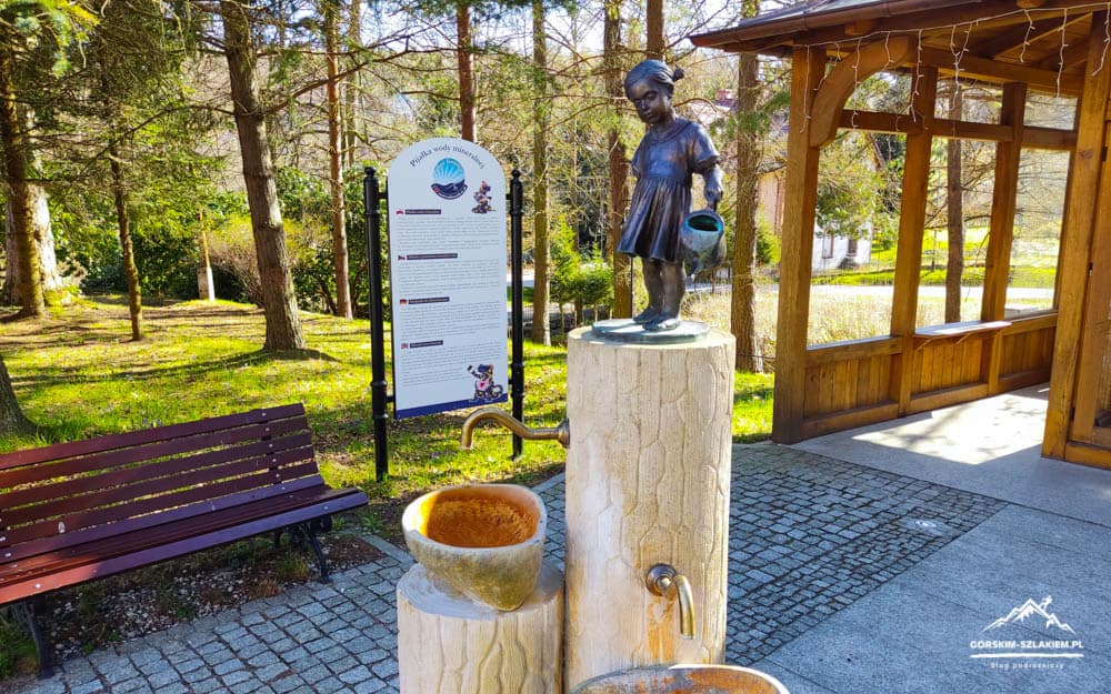 Fontanna z figurą dziewczynki na Górskim Szlaku, otoczona zielonym lasem i miejscem do odpoczynku.