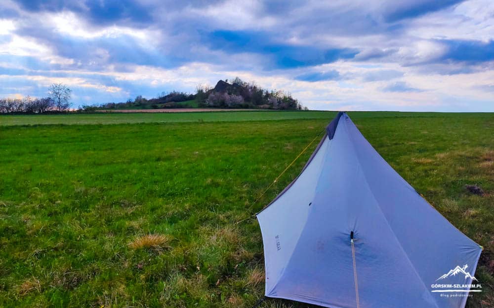A light blue tent pitched on a grassy plain with a distant rocky hill and cloudy sky in the background.
