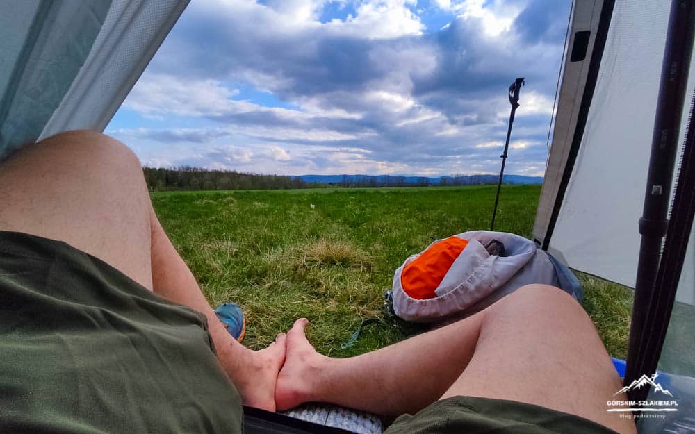 Person resting inside a tent with legs stretched out, overlooking a grassy field under a cloudy sky, with a backpack nearby.