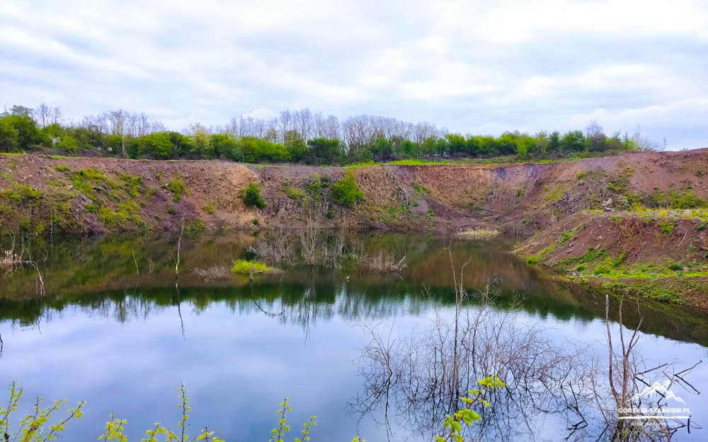 Calm crater lake surrounded by eroded brown cliffs and green vegetation, reflecting a cloudy sky in the water