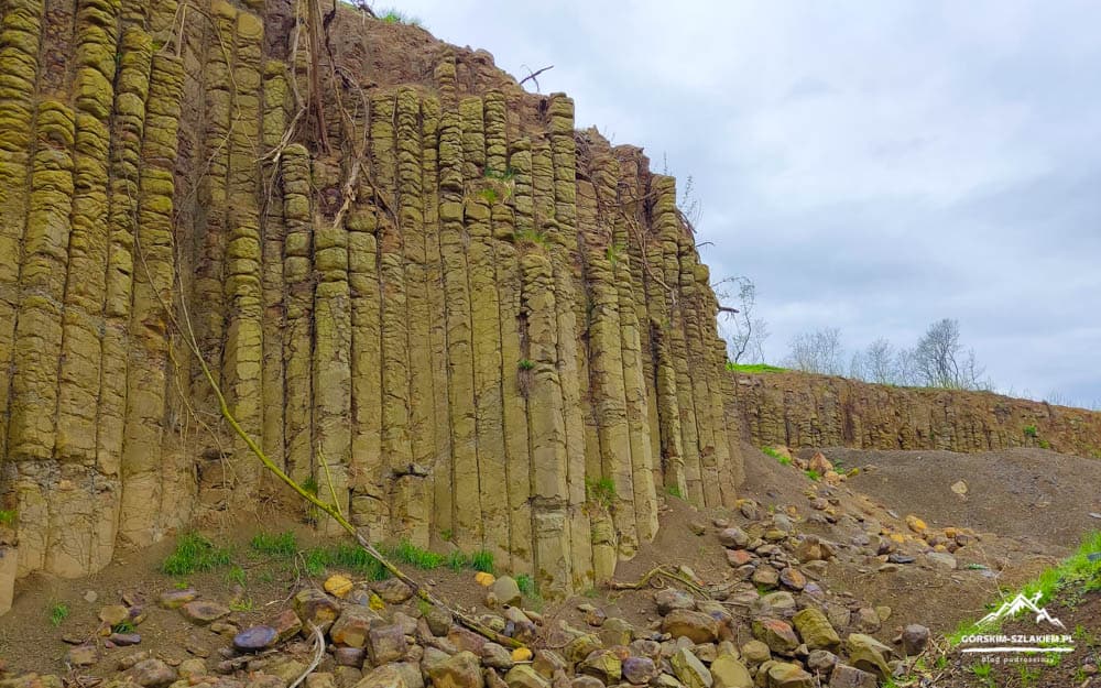 Vertical basalt columns forming a cliff at a rocky quarry, with scattered rocks and sparse vegetation under a cloudy sky.