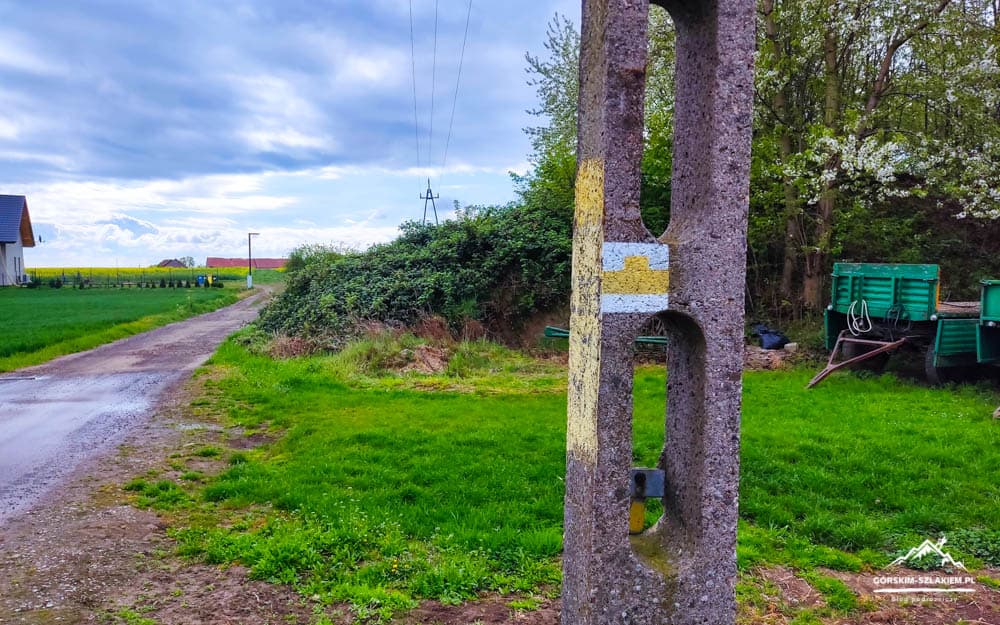 Rural scene with a weathered concrete utility pole in the foreground, a muddy dirt road to the left, and green fields and trees on both sides. A green farm trailer sits among trees on the right, with power lines overhead and a cloudy sky above.