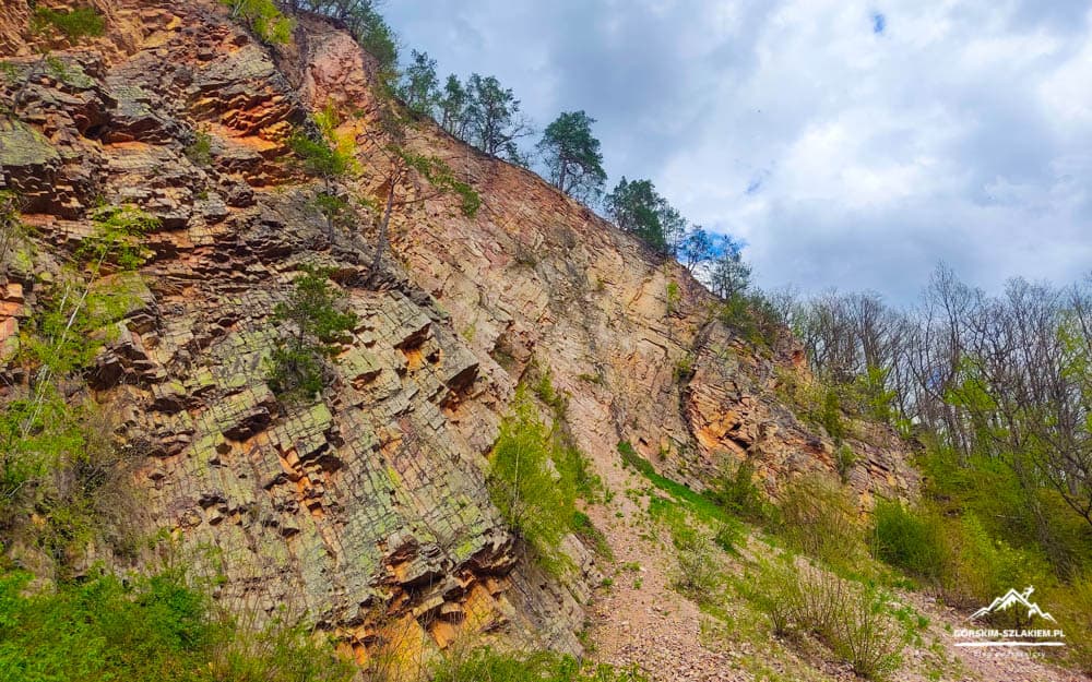 Steep, layered rock cliff with a dirt trail winding uphill and scattered green shrubs under a cloudy sky on a hiking path.