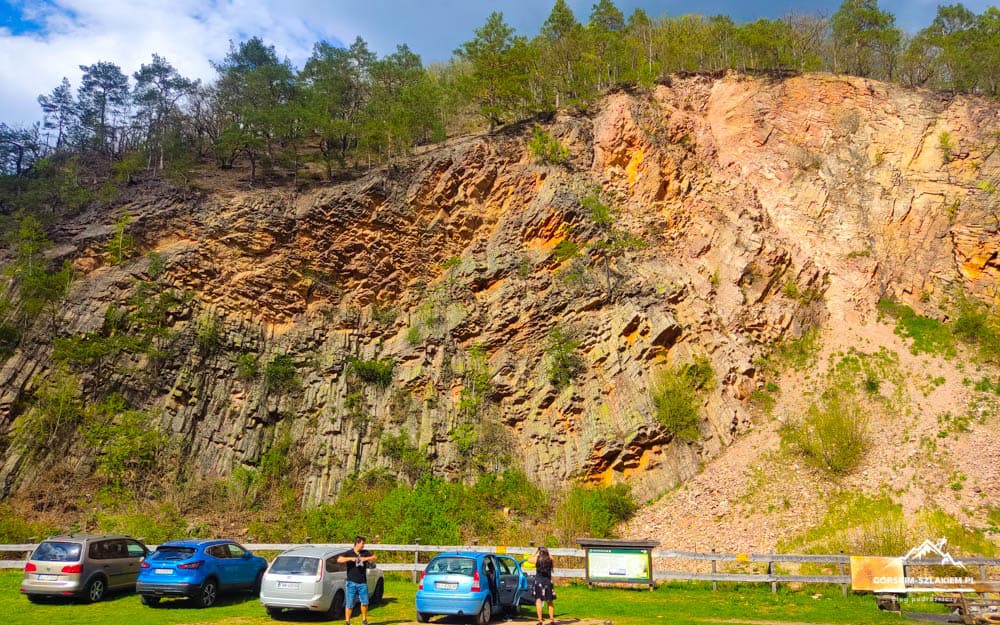 Cliff face with exposed orange-brown rock rises above a parking area where several cars are parked and a few people stand nearby.