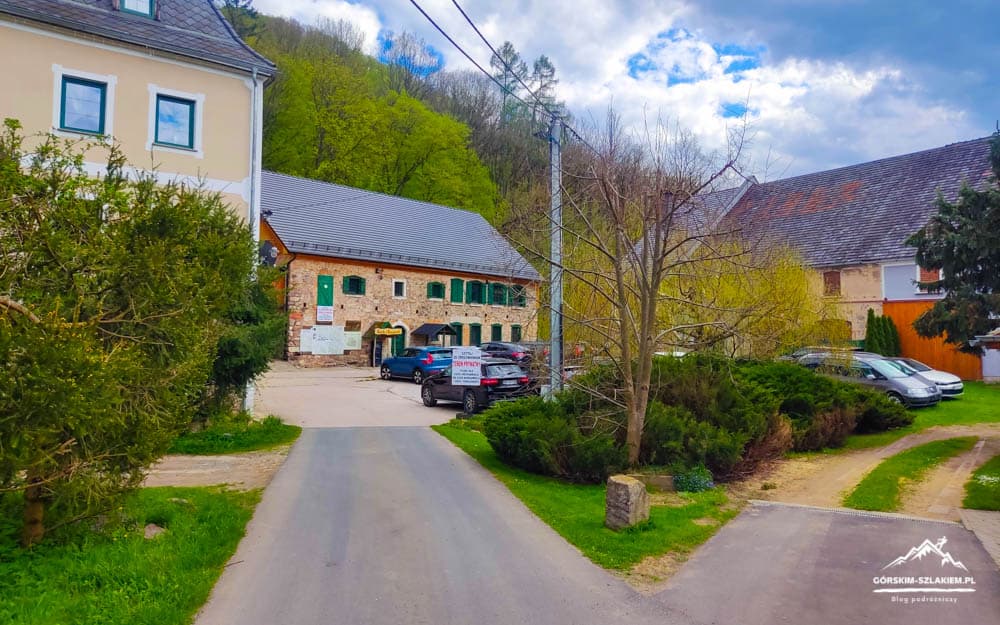 Quiet village street scene with a stone building, green shutters, and several parked cars, surrounded by trees and shrubs under a partly cloudy sky; a small road curves to the right and a gravel path runs beside it.