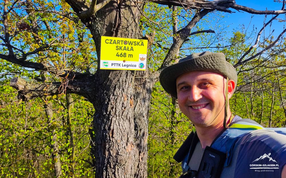 Hiker in a hat and backpack posing beside a yellow trail sign on a tree reading Czartowska Skała 468 m (PTTK Legnica).