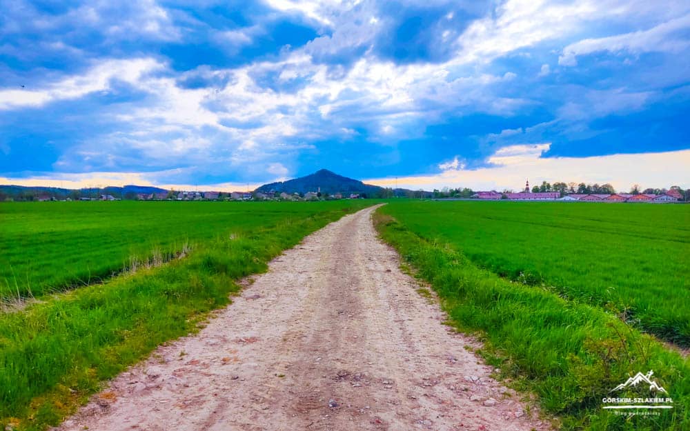 Dusty dirt road cuts through green fields toward a distant village under a dramatic blue-sky with clouds.