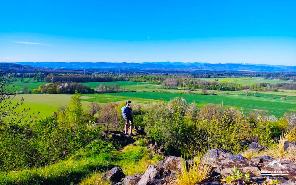 Hiker with backpack standing on a rocky overlook, looking over green fields and distant hills under a bright blue sky.