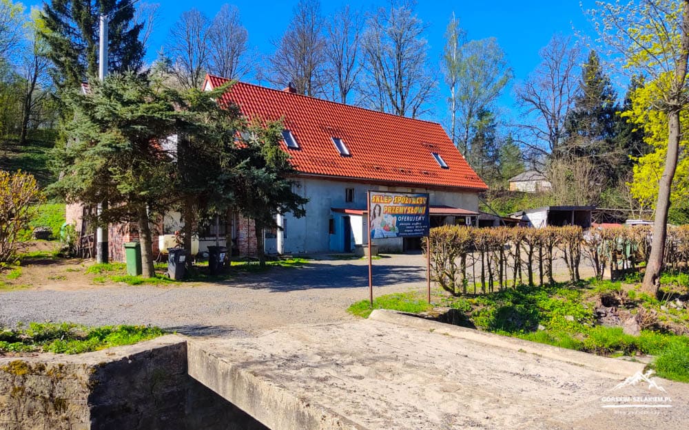 Country house with a red-tiled roof and a sign in front, surrounded by trees and a gravel path on a sunny day from a slight distance.