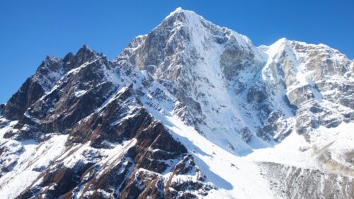 Snow-covered jagged mountain peaks rise against a bright blue sky in a high-alpine landscape.