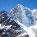 Snow-covered jagged mountain peaks rise against a bright blue sky in a high-alpine landscape.