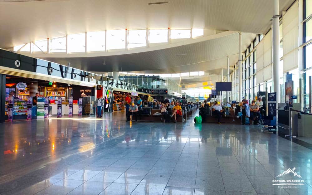 Bright airport terminal with shops along the left, travelers seated across the center, and floor-to-ceiling windows on the right side.