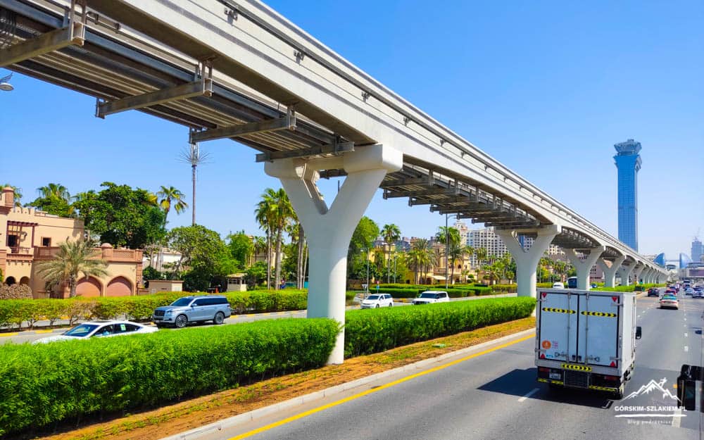 Elevated rail tracks run above a busy highway with palm trees and a white support pillar under a bright blue sky.
