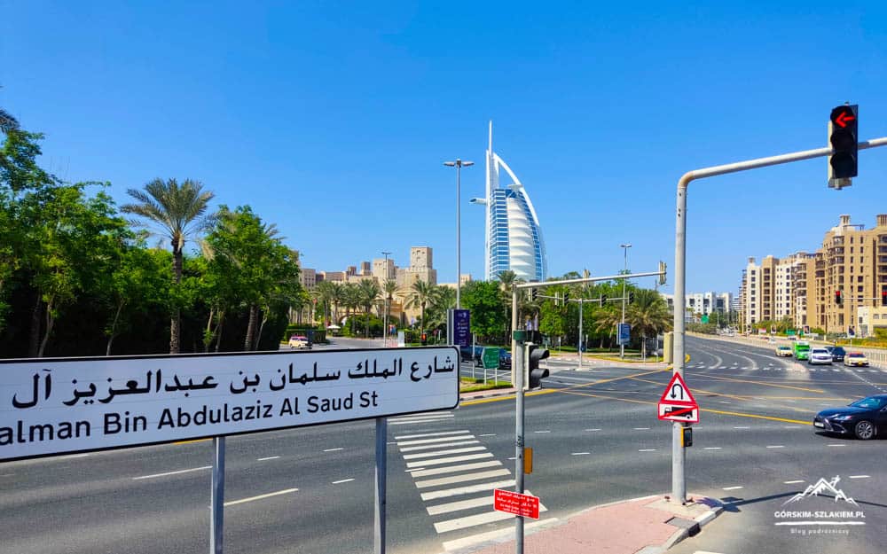 Dubai street scene with Burj Al Arab in the distance and a bilingual street sign in the foreground (Arabic and English).