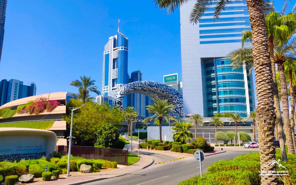 Wide-angle view of a sunlit urban district with tall glass skyscrapers, palm trees, and a large black-and-white patterned arch sculpture by the road.