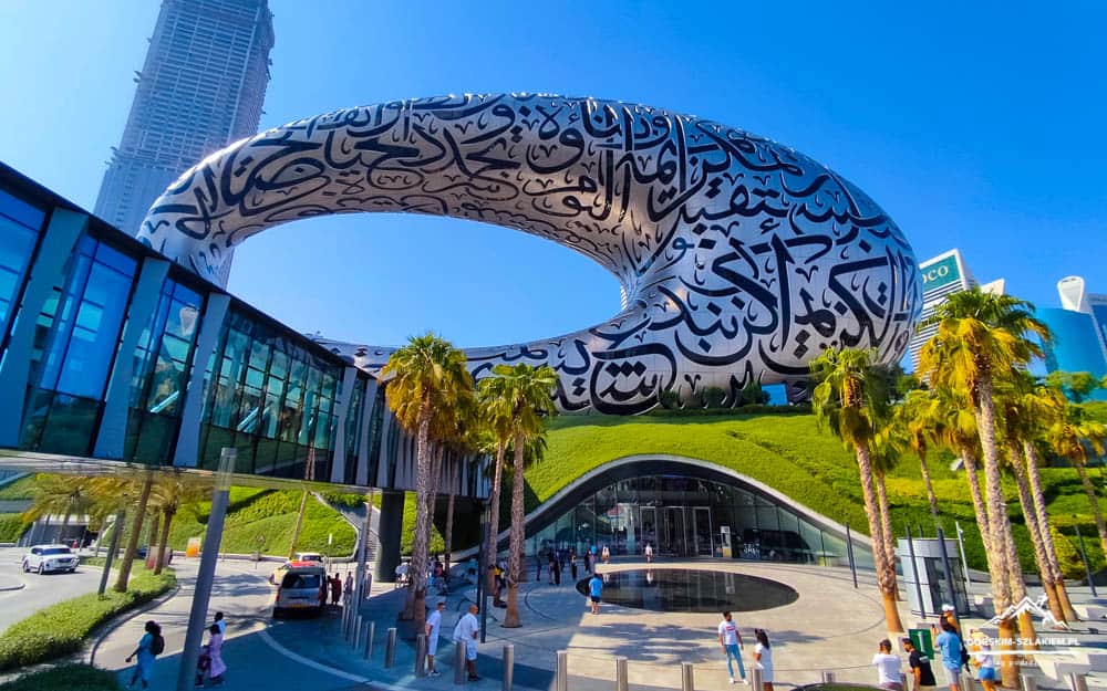 Dubai's Museum of the Future: a white torus-shaped building with black Arabic calligraphy, set against a blue sky with palm trees and a glass walkway.