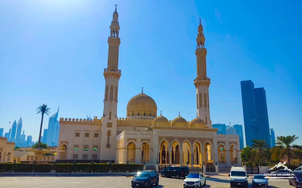 White mosque with gold domes and two minarets in a parking lot, city skyscrapers in the background.
