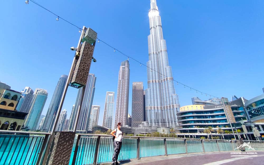 Person poses on a waterfront promenade with Dubai's skyline and Burj Khalifa in the background.