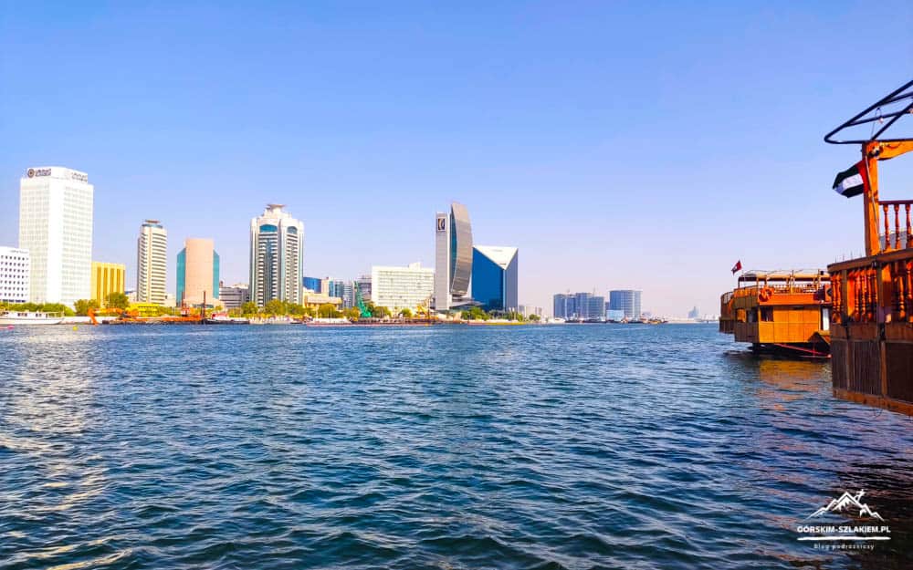 Wide harbor scene with a calm blue body of water and a city skyline of tall buildings along the shore, plus wooden boats docked on the right.