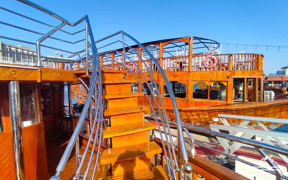 Wooden-decked boat with curved metal staircase and polished railings on a sunny day by the water.