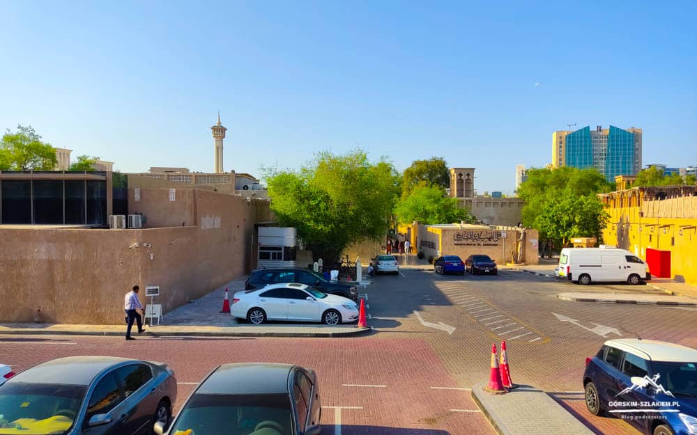 Urban street scene with parked cars along beige walls, trees, and a mix of traditional and modern buildings under a clear blue sky.