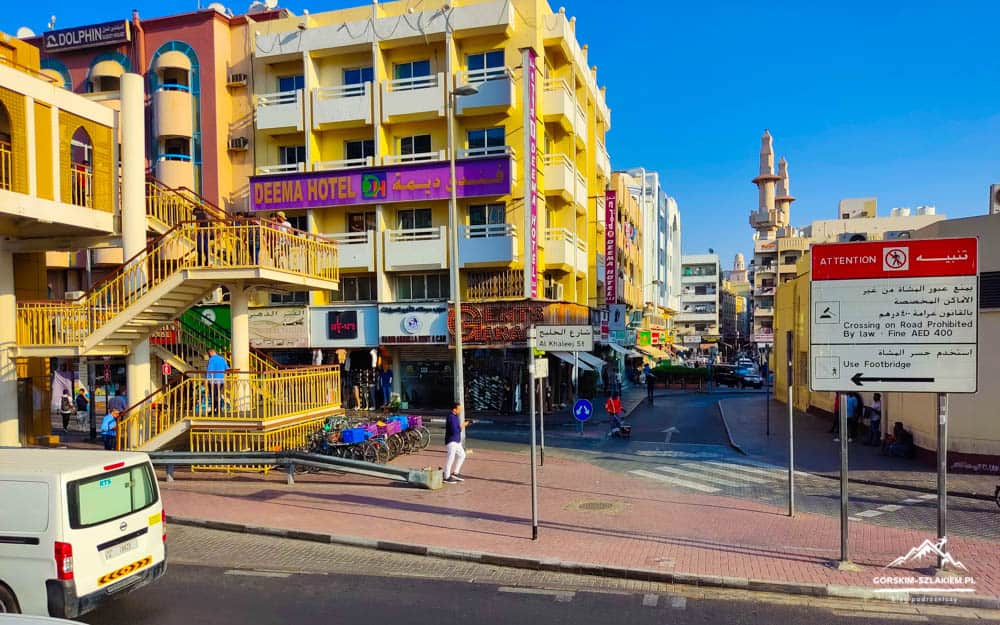 Busy street scene with a pastel yellow building, external staircase, pedestrians, bicycles, and a large signboard; 'Deema Hotel' sign visible against a blue sky