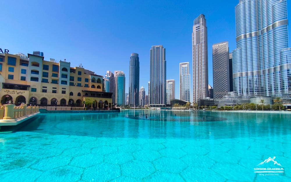 Turquoise pool in the foreground with a row of beige arch-front buildings and a skyline of tall glass skyscrapers under a clear blue sky.