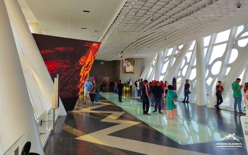 Public gallery hallway with geometric floor pattern and white angled supports; people walk and chat near a large red LED sculpture on the left.