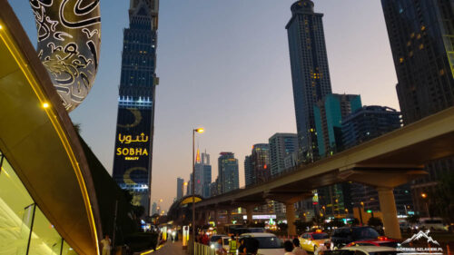 Dubai skyline at dusk featuring the Sobha Realty tower, a raised monorail/rail line, and a busy street with cars and pedestrians nearby