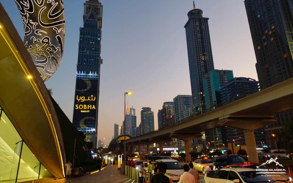 Dubai skyline at dusk featuring the Sobha Realty tower, a raised monorail/rail line, and a busy street with cars and pedestrians nearby