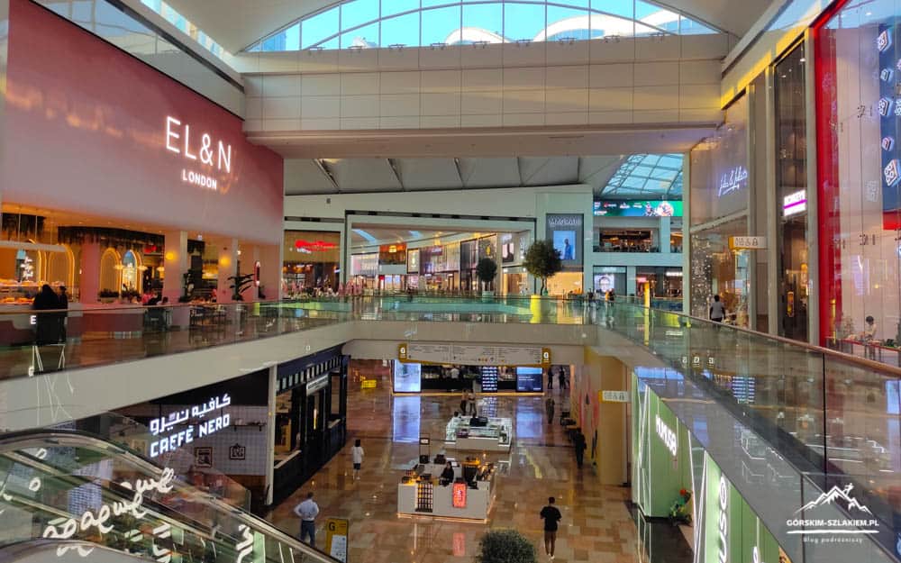 Interior view of a multi-level shopping mall with glass railings, a bright PE storefront for EL&N London, and shoppers on both levels under a skylit atrium.