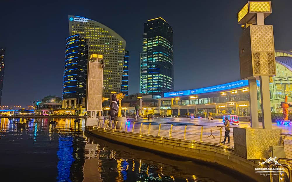 Night city waterfront at dusk with illuminated modern buildings and lampposts along a quay, reflecting in calm water.