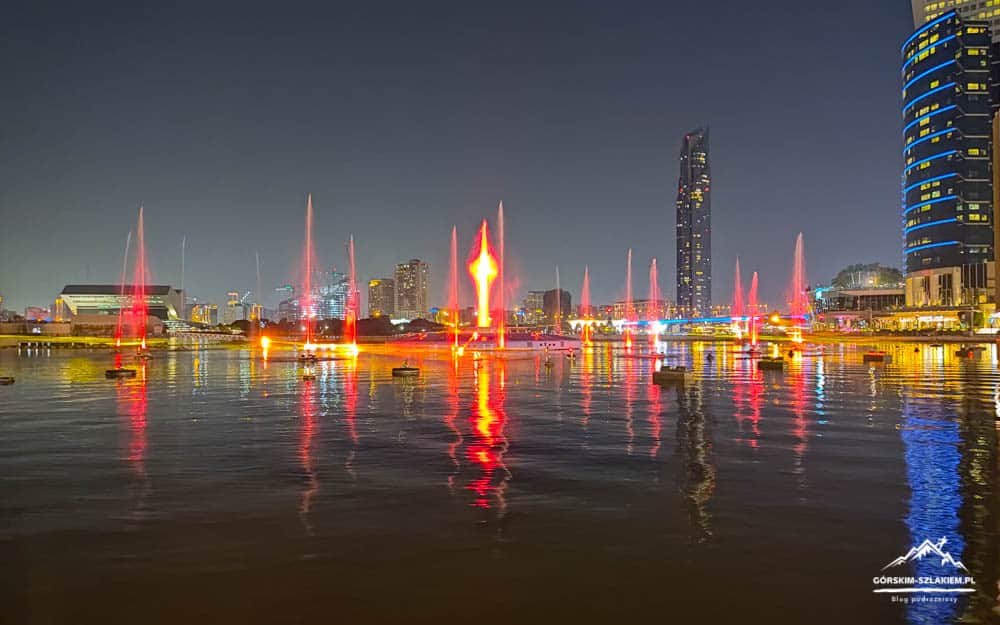 Nighttime harbor scene with red fountain lights spraying from the water and reflecting on the calm surface, city skyline in the background.