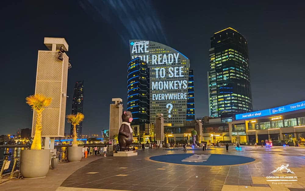 Night view of a city square by the water with a curved skyscraper displaying 'ARE READY TO SEE MONKEYS EVERYWHERE?'. A large bear statue stands in the foreground.