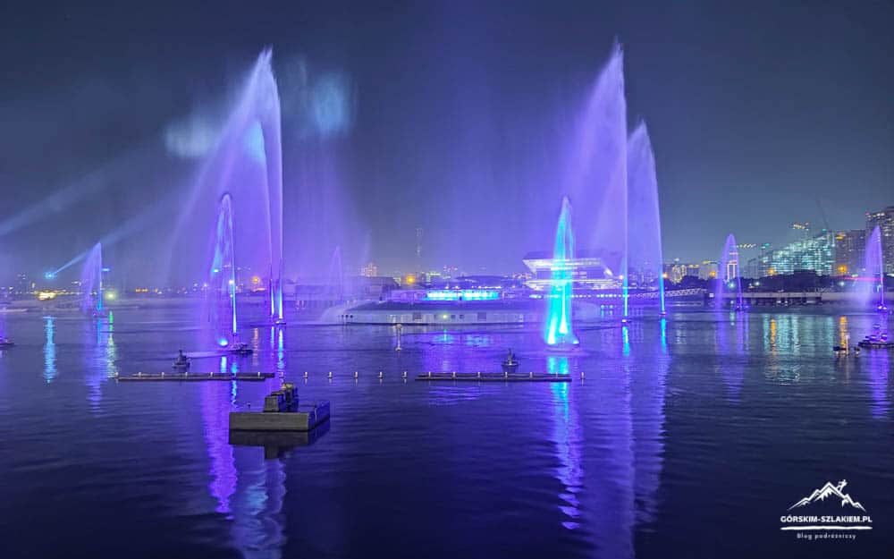 Night harbor fountain show with tall blue and purple water jets, reflected in calm water and city lights in the distance.