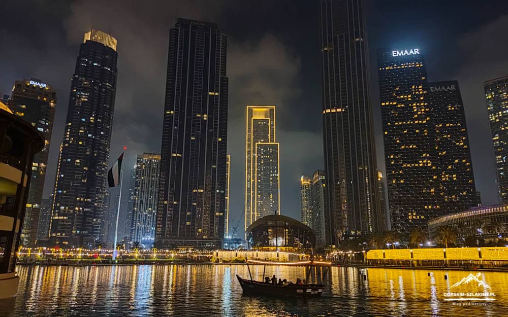 Nighttime cityscape with illuminated skyscrapers reflecting on the water and a small boat in the foreground.
