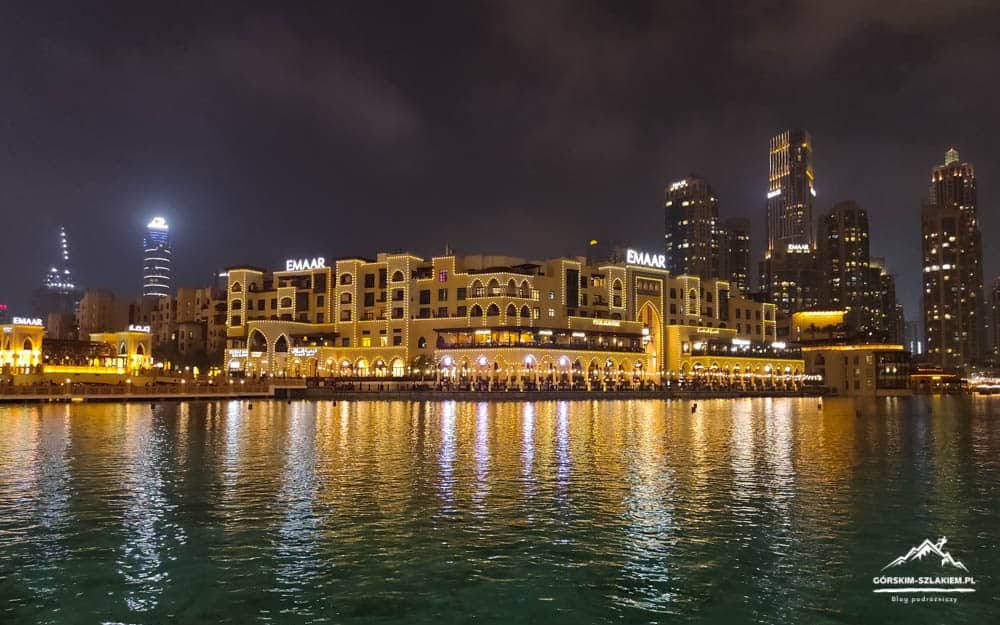 Nighttime waterfront cityscape with illuminated arches and beige buildings reflecting on calm water.