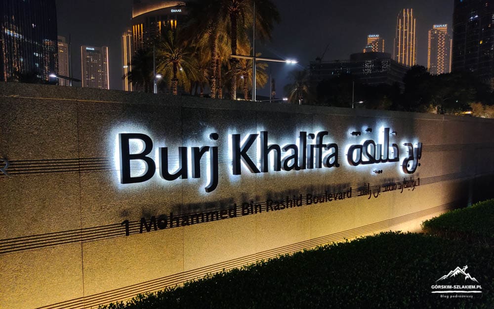 Night view of a stone wall with illuminated 'Burj Khalifa' sign and palm trees, with Dubai skyline in the background at night.
