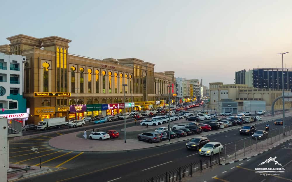 Busy urban street scene with a large beige mall building and arched windows, storefronts glowing at dusk and a line of cars along the road near a roundabout.