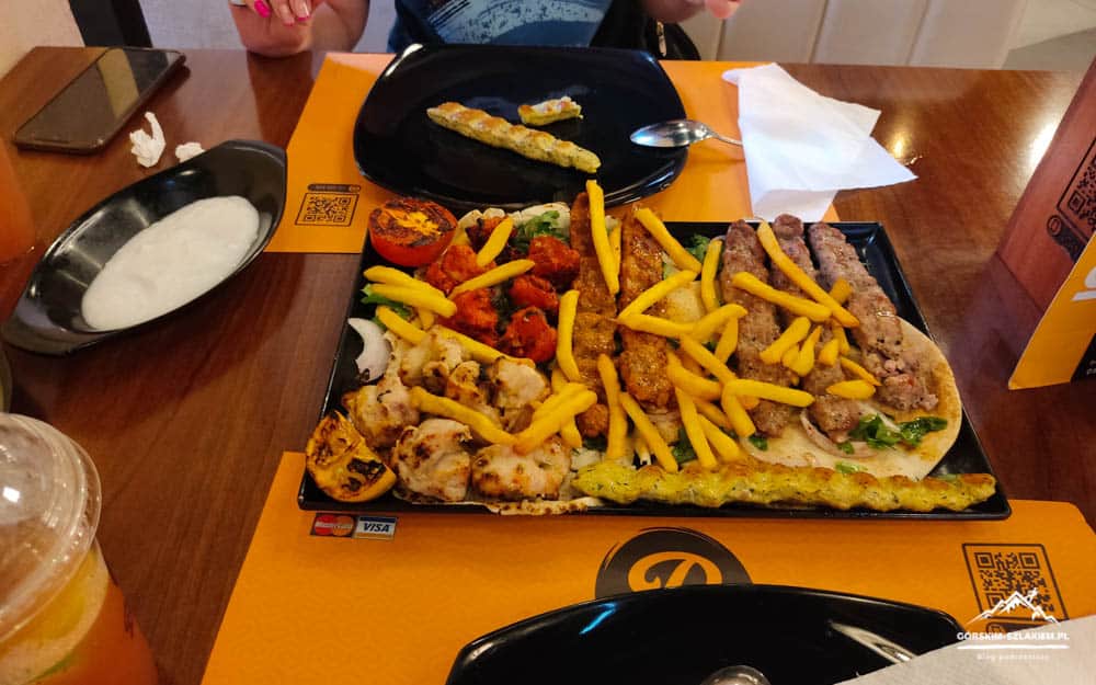 Large tray of mixed grilled meats with fries and vegetables on a table at a restaurant, with a bowl of white sauce nearby.
