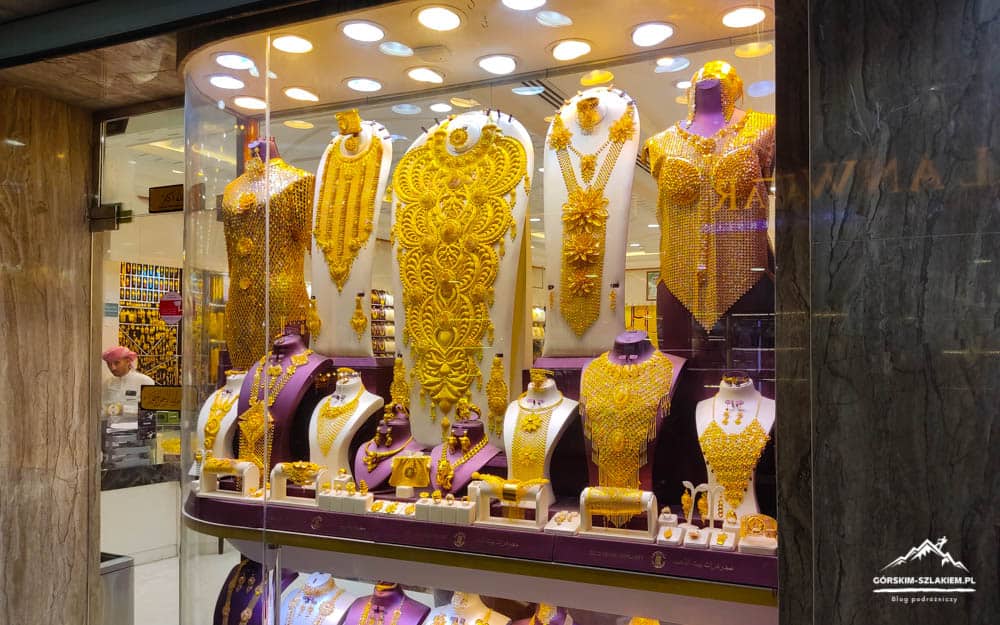 Gold jewelry display in a shop window with ornate necklaces and bracelets on purple stands under bright lights.