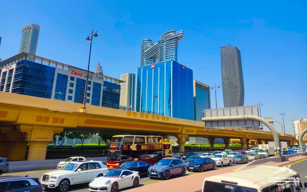 Busy city street with a yellow elevated railway track and modern blue glass skyscrapers in the background.