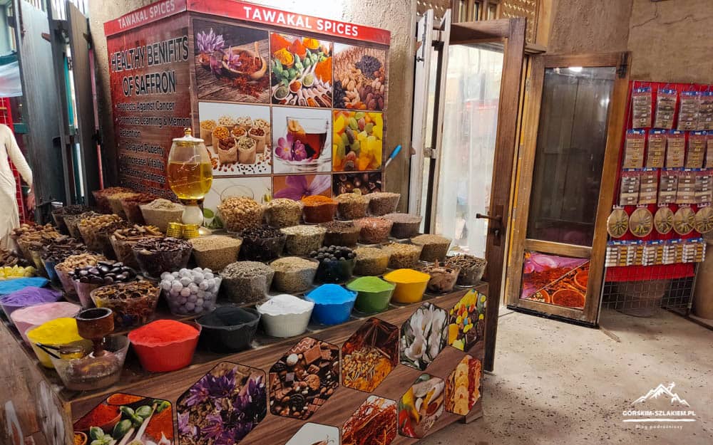 Spice stall with colorful bowls of powders and spices on a wooden display, storefront sign 'TAWAKAL SPICES' in the background